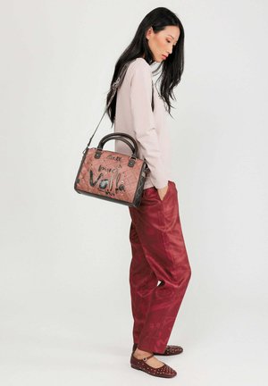 Borsa a tracolla rosa trapuntata con dettagli neri e hardware metallico, caratterizzata da testo ricamato. Il modello indossa un top beige e pantaloni rossi.