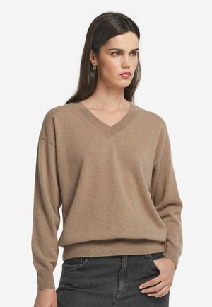 Sweter z dekoltem w kształcie V, z długim rękawem, w miękkim beżowym materiale; luźny krój z prążkowanym dołem i mankietami; minimalna tekstura i brak widocznych wzorów.