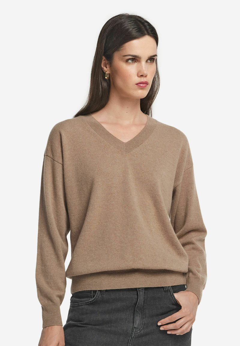 V-Ausschnitt, langärmliger Pullover aus weichem beigen Material; lockere Passform mit geripptem Saum und Bündchen; minimale Textur und keine sichtbaren Muster.
