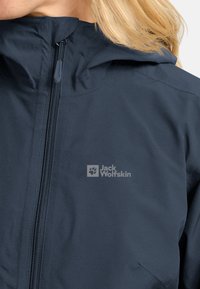 Giacca impermeabile blu marina con colletto alto, zip completa e logo sul lato sinistro. Texture liscia con design leggero.