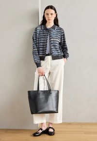 Strikket cardigan i blå og grå mønster med sort krave, parret med lyse bredbenede bukser og en sort tote bag. Sorte sko fuldender looket.