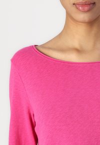 Rosa långärmad topp med bred halsringning, som har en slät textur och en figurnära silhuett, tillverkad av ett mjukt, lätt tyg.