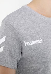 Camiseta de algodón gris con mangas cortas, presenta el logotipo blanco "hummel" en el pecho y chevrones blancos en la manga izquierda. Textura suave.