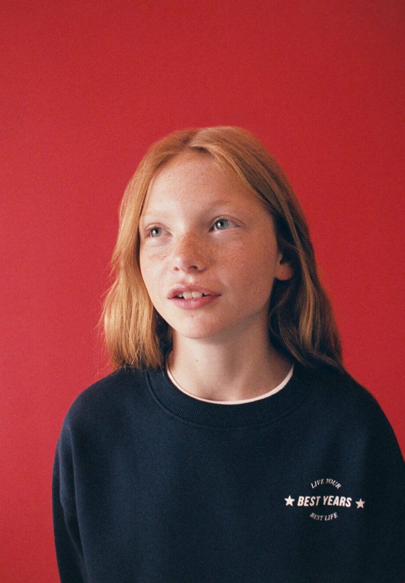 Jeune personne rousse avec des taches de rousseur portant un sweat-shirt sombre avec le texte "Best Years" sur un fond rouge, regardant légèrement vers le haut.