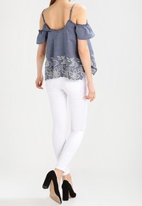 Top off-shoulder in chambray blu con maniche a volant, ricamo floreale bianco sull'orlo, abbinato a jeans skinny bianchi e tacchi alti neri.