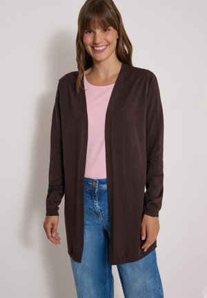 Femme souriante portant un long cardigan marron foncé ouvert sur une chemise rose clair et un jean bleu, debout devant un fond uni.