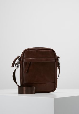 Compact donkerbruine leren crossbodytas met voorvakje met rits en verstelbare stoffen schouderriem, gepresenteerd op een witte ondergrond.