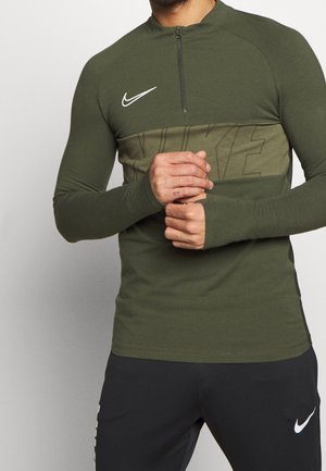 Mężczyzna ubrany w oliwkowozielony top Nike z długim rękawem i zamkiem do ćwiartki oraz czarne spodnie Nike, trzymający ręce splecione na talii na szarym tle.