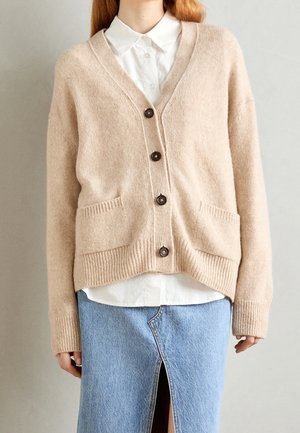 Personne portant un cardigan beige boutonné sur une chemise blanche et une jupe en denim bleu clair avec une fente à l'avant.