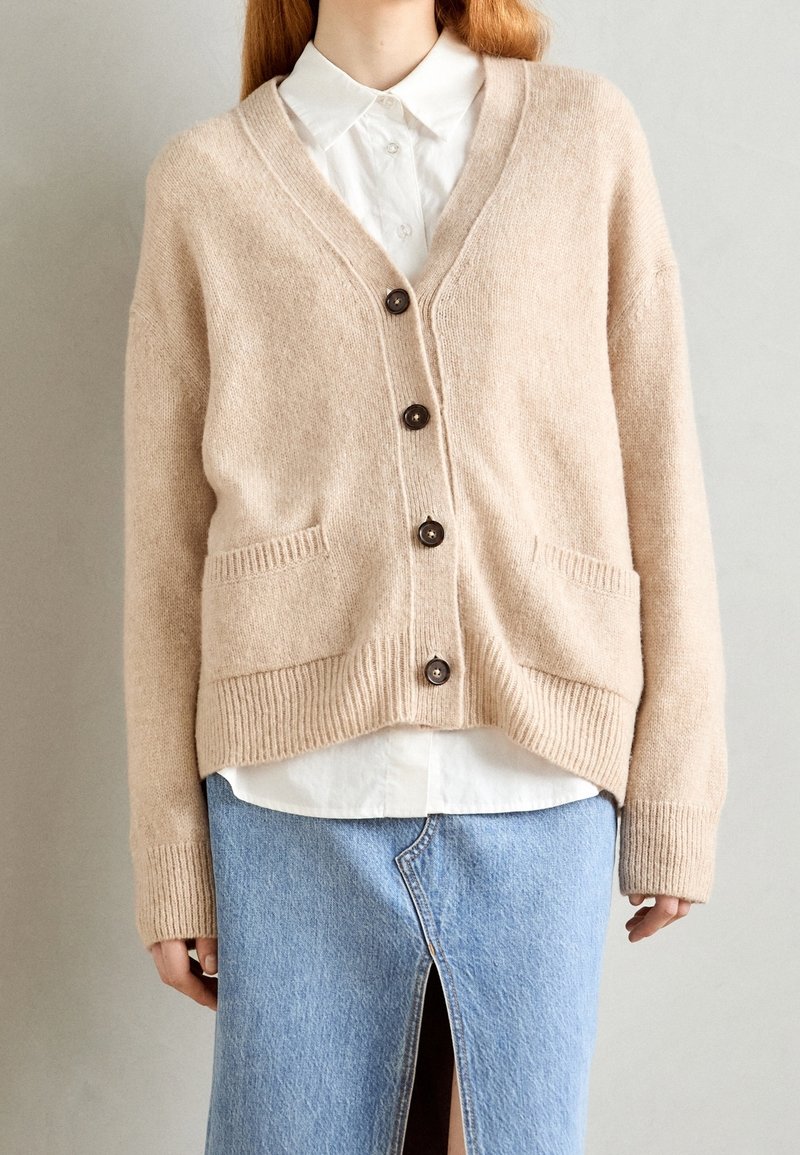 Personne portant un cardigan beige boutonné sur une chemise blanche et une jupe en denim bleu clair avec une fente à l'avant.