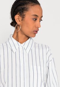 Camicia bianca a righe con colletto, presenta sottili righe verticali blu e una tasca sul petto. Tessuto morbido.