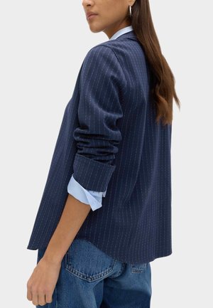 Vrouw met een marineblauw colbert met krijtstreepjes en opgerolde mouwen over een lichtblauw overhemd en een blauwe spijkerbroek, zijaanzicht en achterkant.