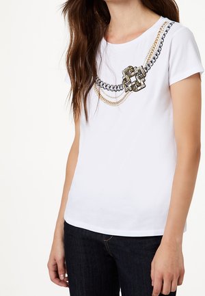 Mujer que lleva una camiseta blanca con un diseño impreso de cadena y cierre en dorado y negro a lo largo del pecho, combinada con vaqueros oscuros.