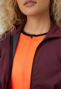 Žena s kučeravými blond vlasmi oblečená v marónovej bunde cez jasne oranžový textúrovaný top s predným zipsom.