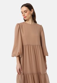 Robe beige clair en tissu texturé. Présente un col rond, des manches ballon et une jupe fluide à volants. Design minimaliste.