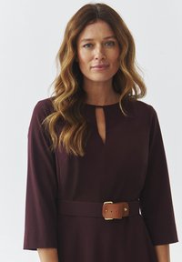 Robe bordeaux avec des manches trois-quarts, décolleté en petits œillets et une ceinture marron avec une boucle en métal. Texture lisse, peu de détails.