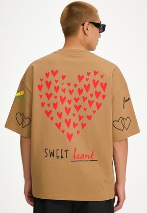 Person trägt ein beige T-Shirt mit roten Herzen, die ein großes Herz bilden, und dem Text "SWEET heart" auf dem Rücken, schwarze Shorts und schwarze Sonnenbrille.