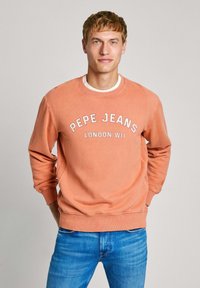 Sudadera de cuello redondo de Pepe Jeans en coral deslavado, con texto blanco "PEPE JEANS LONDON W11" y puños, cuello y dobladillo acanalados. Combinada con jeans azules.