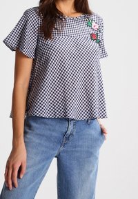 Marinblå och vit rutmönstrad t-shirt med blommig broderad detalj. Flödande, lös passform. I kombination med ljusblå denimbyxor.