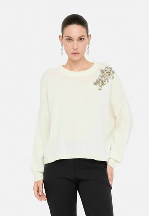 Maglione bianco lavorato a maglia con scollatura rotonda, spalle a caduta e decorazioni di perline sulla spalla sinistra. Abbinato a pantaloni neri.