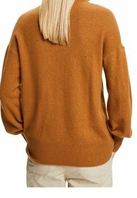 Personne blonde portant un pull brun ample à manches longues et un pantalon beige clair, vue de dos.