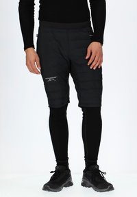 Zwarte gewatteerde shorts met een zachte textuur, voorzien van een logo en een getailleerd ontwerp, gedragen over zwarte leggings en gecombineerd met zwarte schoenen.