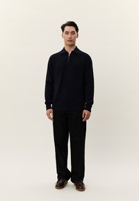 Νέος άνδρας που στέκεται, φοράει σκοτεινό πλεκτό πουλόβερ με φερμουάρ, φαρδιά μαύρη παντελόνια και καφέ δερμάτινα παπούτσια, μπροστά σε απλό φωτει�νό φόντο.