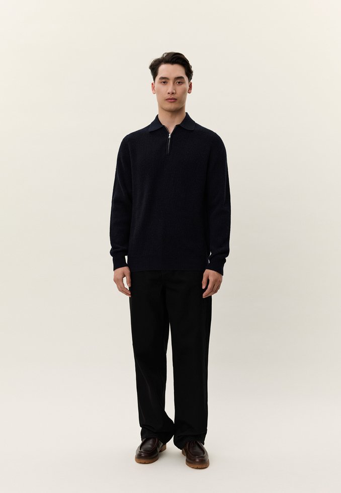 Νέος άνδρας που στέκεται, φοράει σκοτεινό πλεκτό πουλόβερ με φερμουάρ, φαρδιά μαύρη παντελόνια και καφέ δερμάτινα παπούτσια, μπροστά σε απλό φωτεινό φόντο.