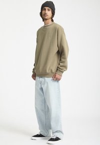Sweat-shirt beige avec des poignets côtelés, jean en denim bleu clair à coupe lâche et baskets noires, présentant un design décontracté et des éléments de marque minimalistes.