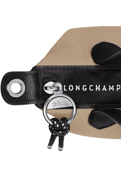 Tan nylon bag with black leather accents, featuring a silver logo plate, zipper pull, and decorative braided cord detail.