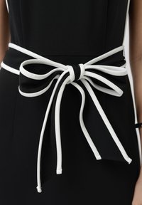 Rochie neagră cu o curea lată în talie legată într-un arc, cu garnitură albă de-a lungul marginilor și capete lungi și largi care atârnă în jos.