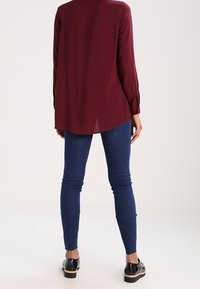 Persona in piedi di spalle che indossa una maglietta a maniche lunghe bordeaux, jeans skinny blu e scarpe con plateau nere su uno sfondo bianco.