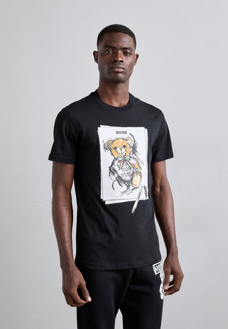 T-shirt en coton noir ornée d'une illustration d'ours avec un nœud papillon et du texte, manches courtes, col rond, portée avec un pantalon noir.
