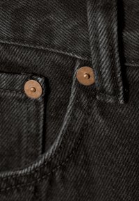 Tecido de denim negro com rebites metálicos de color latón en el bolsillo delantero, con costuras visibles y una textura suave.