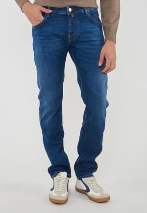 Herren Jeans aus blauen Denim mit Slim Fit, Kontrastnähten, fünf Taschen und einem Knopfverschluss. Getragen mit weißen und grauen Sneakers.