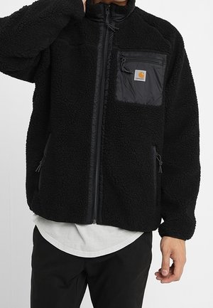 Zwart fleecejack met ritssluiting en gestructureerde stof, borstzak met Carhartt-logo, gedragen over een wit shirt en zwarte broek.