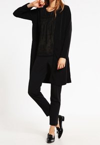 Mujer vistiendo un cárdigan largo negro, top negro transparente, pantalones ajustados negros y mocasines de charol negros, de pie con una pierna cruzada.