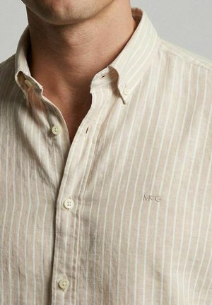 Mann trägt beige Hemd mit weißen vertikalen Streifen und kleinem gesticktem "MCG"-Logo auf der Brust.