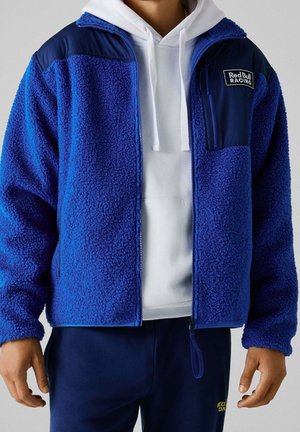 Man wearing a blue fleece jacket with Red Bull Racing logo over a white hoodie and navy pants, standing with arms relaxed by sides.