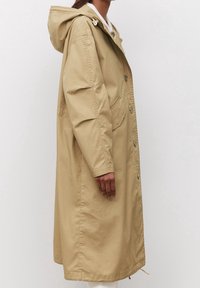 Abrigo impermeable beige con capucha, que cuenta con bolsillos laterales, un cierre de botones y un corte ligeramente holgado. Textura suave, diseño hasta las rodillas.