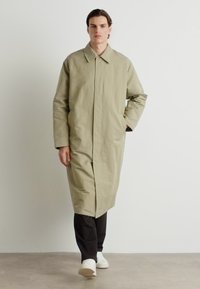 Lichtgroene trenchcoat met een kraag, lange mouwen en een voorkrui, gedragen over een donkere broek en witte sneakers op een neutrale achtergrond.
