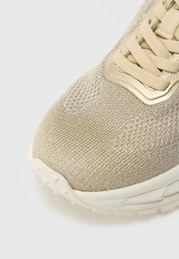 Primo piano della punta di una sneaker beige lavorata a maglia con suola texturizzata e lacci abbinati su sfondo bianco.