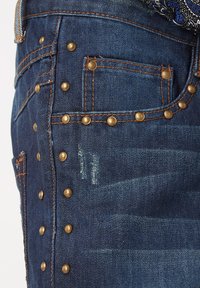 Los jeans de mezclilla oscura cuentan con remaches de latón a lo largo de las costuras laterales y los bolsillos, con un contraste de costuras en naranja y áreas sutilmente desgastadas.