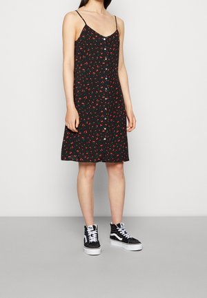 Femme portant une robe noire sans manches avec un petit imprimé floral rouge et des boutons blancs sur le devant, associée à des baskets montantes noires et blanches.