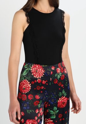 Femme portant un haut noir sans manches avec un détail en dentelle et un pantalon taille haute foncé avec de grandes impressions florales rouges, vertes et bleues.