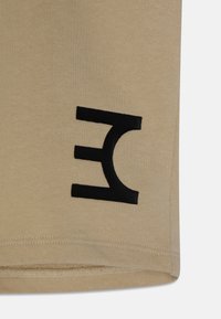 Béžová bavlněná tkanina s hladkou strukturou, na levé straně se nachází výrazné černé tištěné logo. Lem je ušit s čistým zakončením.