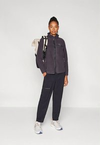 Siva flis jopica z zadrgo spredaj in stranskima žepoma, kombinirana s temnimi hlačami in športnimi copati. Model nosi svetel nahrbtnik.