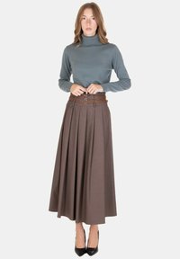 Donna che indossa un maglione grigio a collo alto, gonna midi plissettata marrone con cintura doppia fibbia e tacchi neri a punta, in piedi contro uno sfondo bianco.