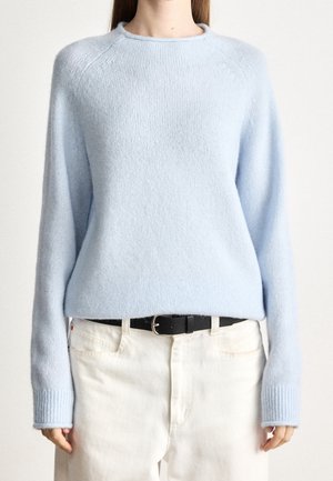 Personne portant un pull tricoté bleu pâle à manches longues rentré dans un pantalon blanc avec une ceinture noire, debout devant un fond uni.