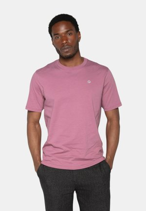 Homme portant un t-shirt simple rose pâle à manches courtes et un pantalon gris foncé, se tenant avec les mains dans les poches devant un fond blanc.